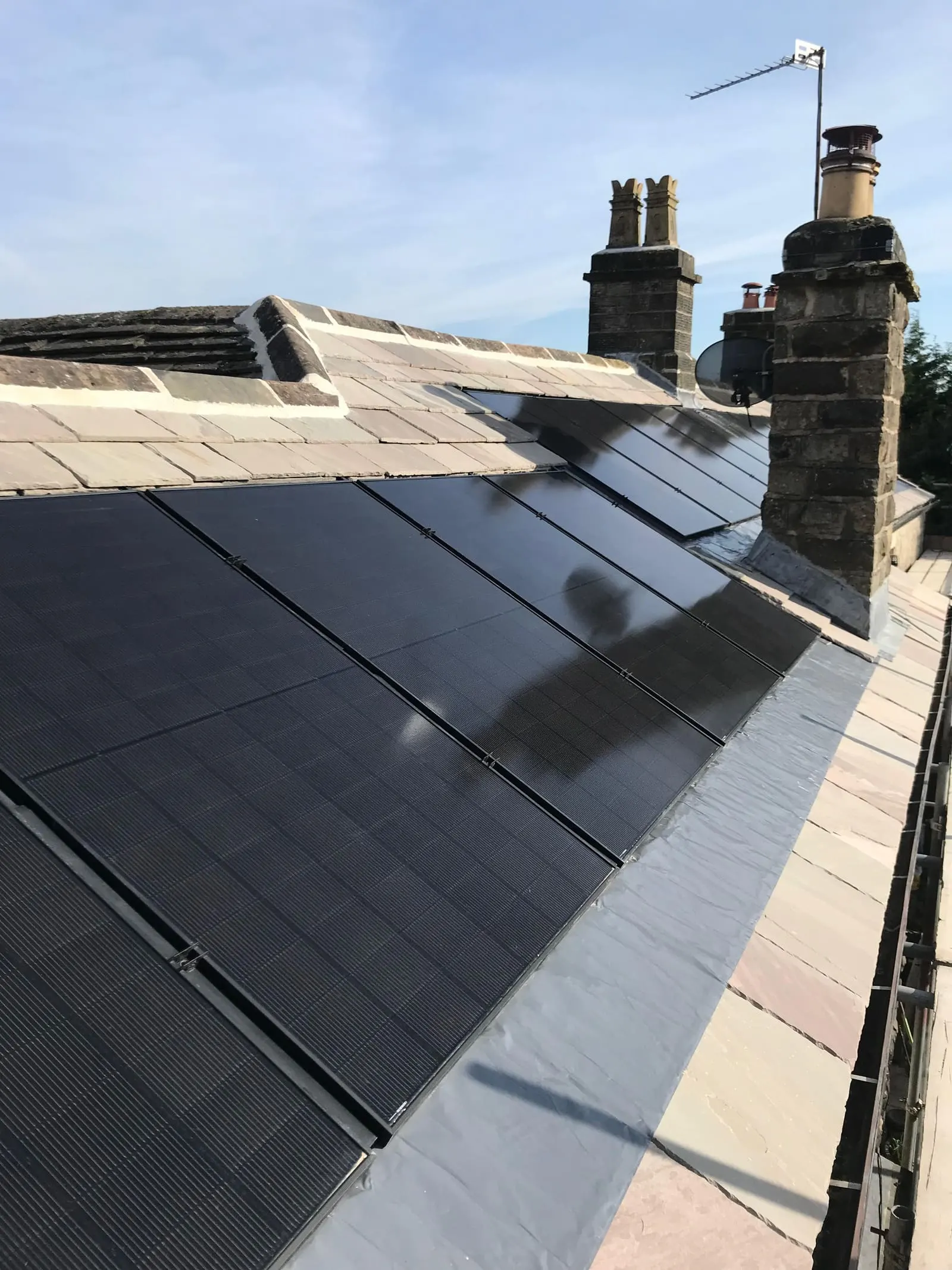All-black solar PV panels on a slate roof, with stone chimney stacks and lead ridge work behind