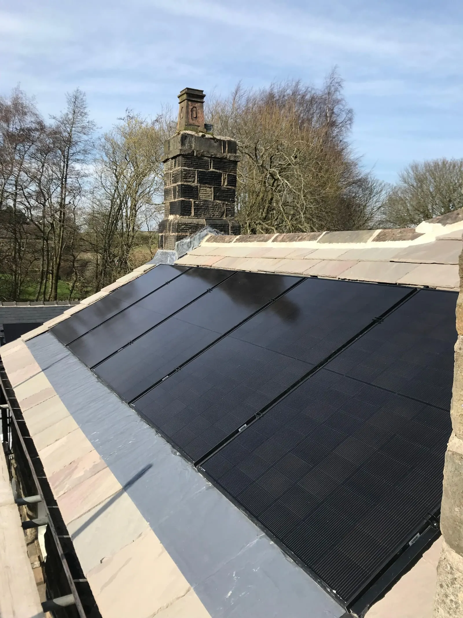 All-black solar panels on a slate roof with a tall stone chimney and open countryside behind