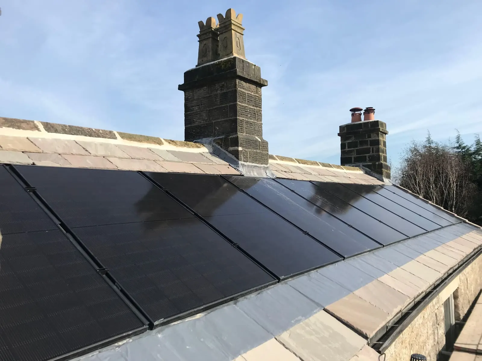 Wide view of the all-black solar array spanning the roof between two stone chimney stacks