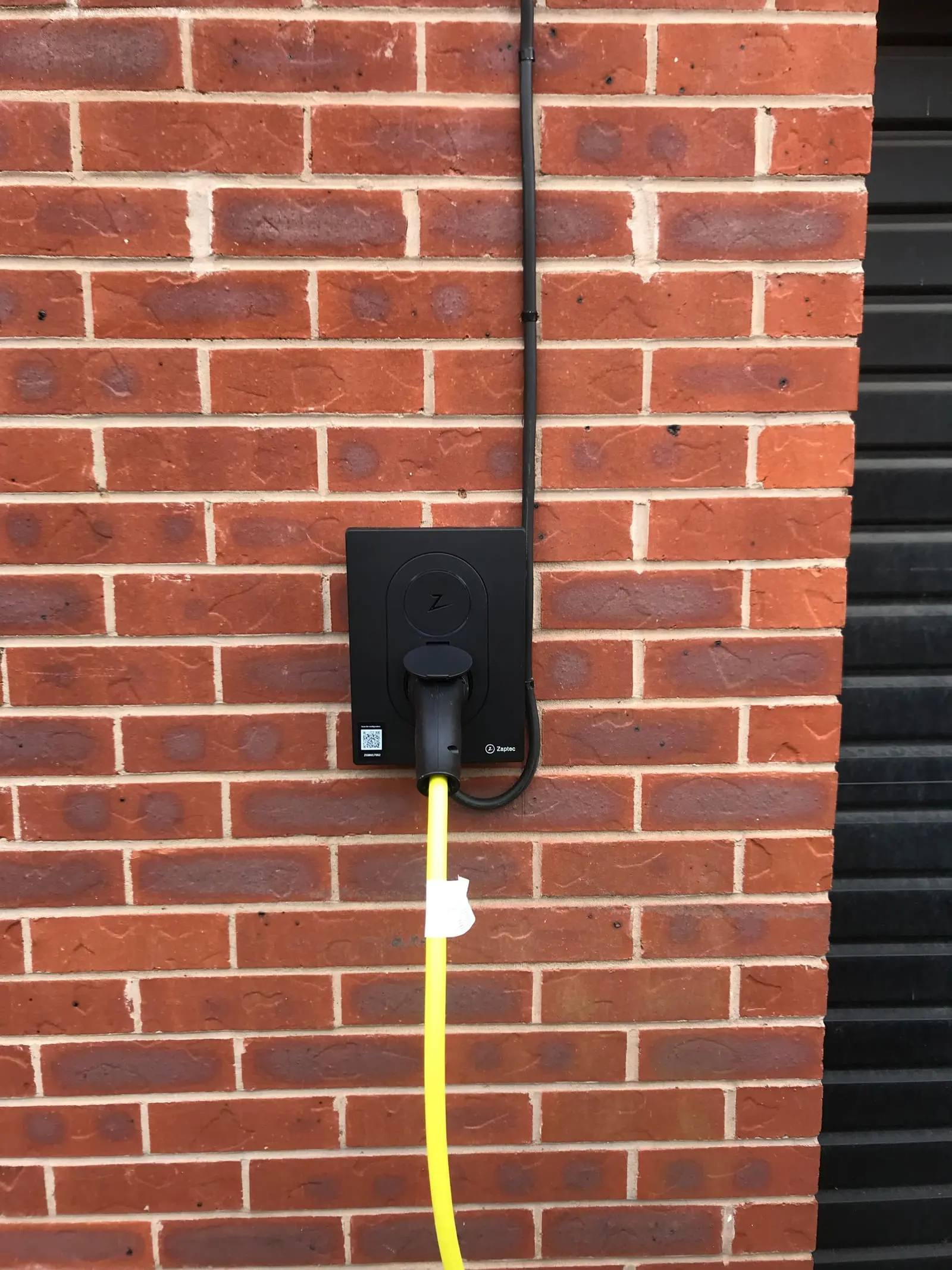 Finished black Zaptec EV charger on a brick wall with a yellow charging cable plugged in