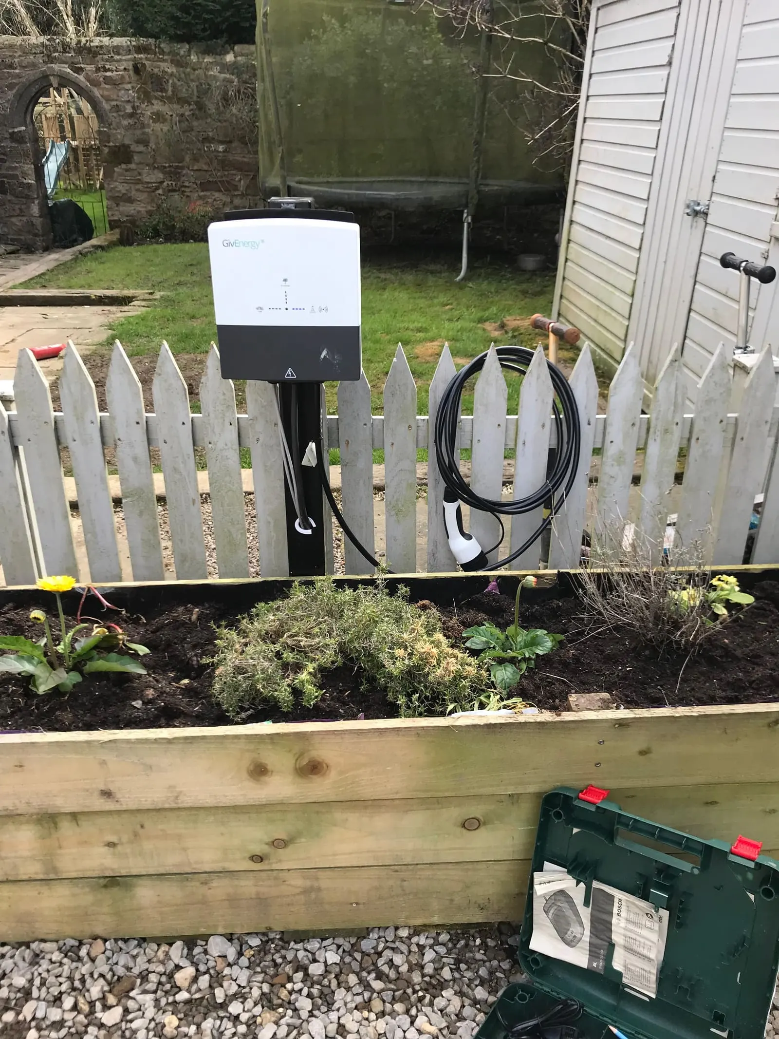 EV charger mounted on a post by a raised flower bed and picket fence, with a coiled tethered charging cable
