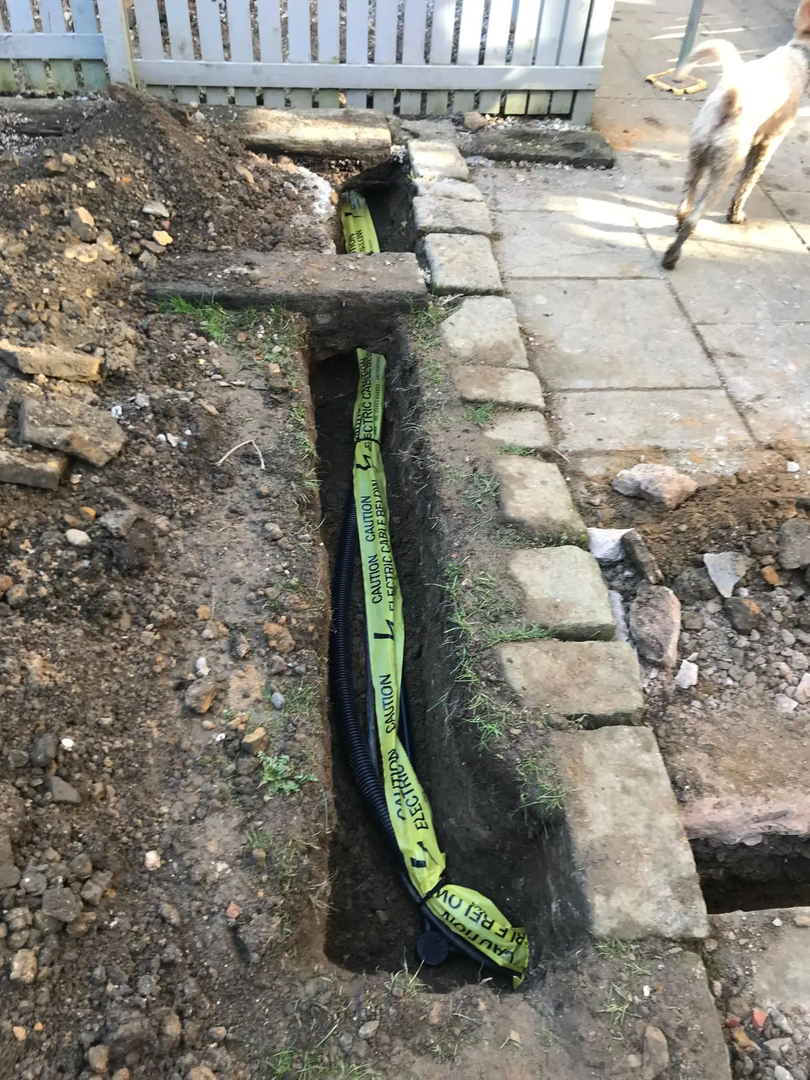 Hand-dug trench across paving with ducted armoured cable and yellow warning tape laid in
