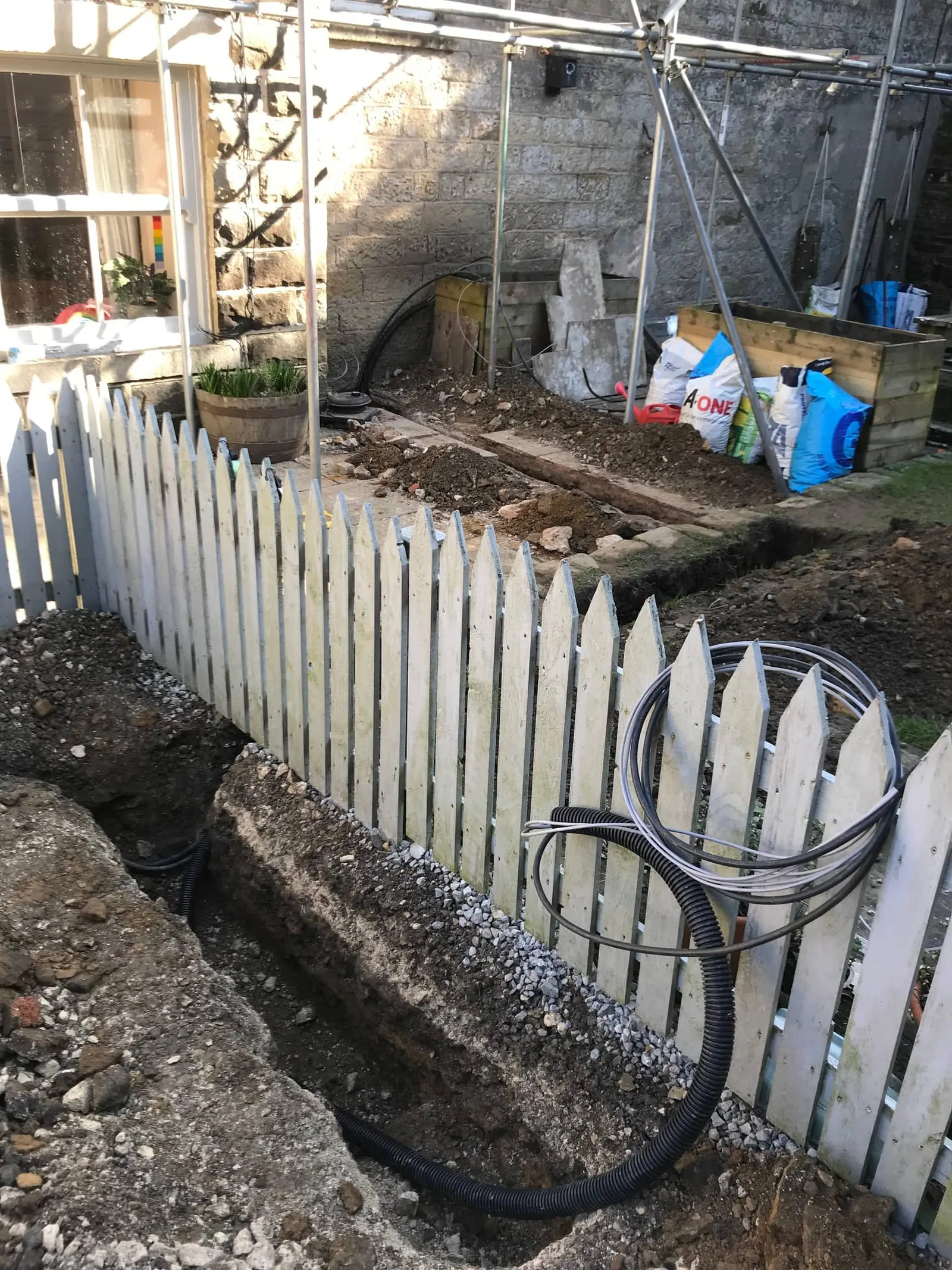 Trench running alongside a white picket fence with coiled cable ducting ready to lay