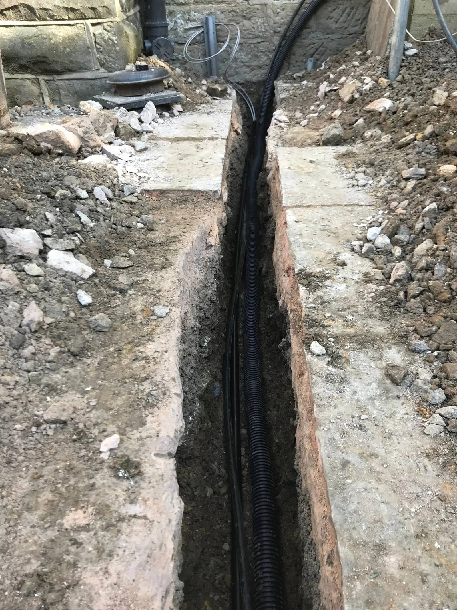 Close-up of the trench between a stone wall and paving, with armoured cable ducting run along the base